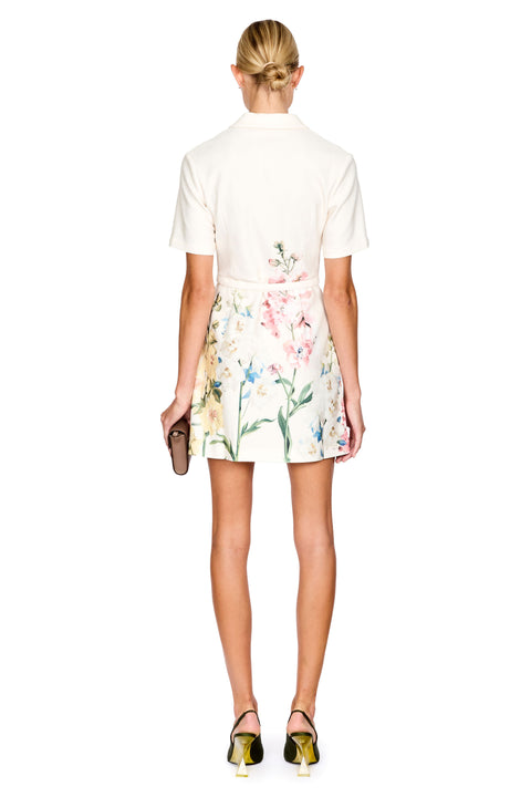 A model shown from the back wearing a cream short-sleeve mini dress with a pink, green, and blue floral print. She is holding a small brown clutch and wearing black and gold high heels against a solid white background.