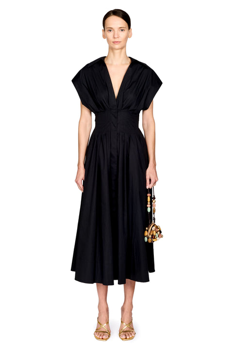 A model wearing a black midi dress with a deep V-neckline, short draped cap sleeves, and a gathered waist, paired with gold sandals and a small beaded handbag against a plain white background.
