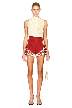 A model stands against a white studio background wearing a sleeveless cream top with a draped tassel detail and red shorts featuring cream floral embroidery along the hem. She is holding a small cream handbag and wearing clear-strap high-heel sandals.
