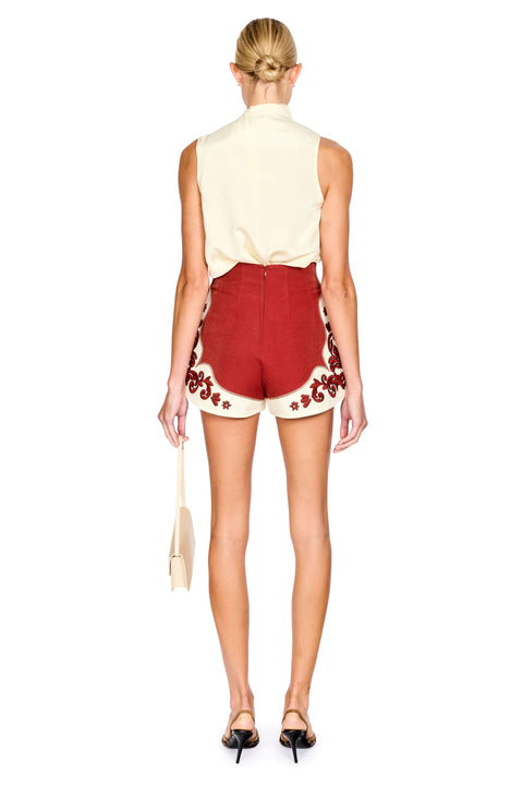 A model shown from the back wearing a cream sleeveless top and red shorts with cream floral embroidery, holding a cream handbag against a white background.