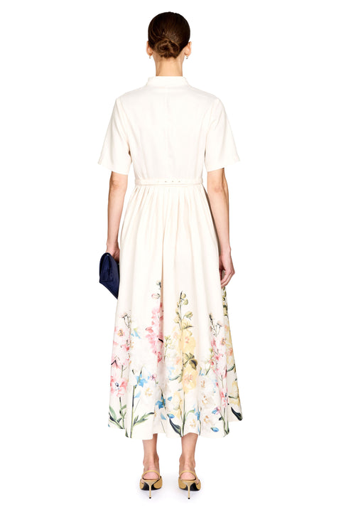 A woman viewed from the back wearing a cream-colored midi dress with a short-sleeve collar, a defined waist, and a colorful floral print concentrated along the lower skirt. She is holding a small dark blue clutch and wearing pointed-toe heels, set against a plain white background.