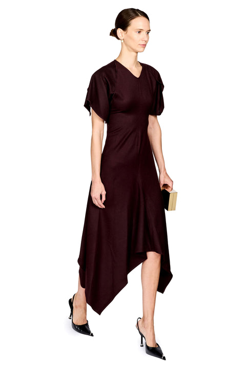 A woman modeling a dark burgundy midi dress with short puff sleeves, a V-neckline, and an asymmetrical draped hem, paired with black pointed-toe heels and holding a small gold clutch against a plain white background.