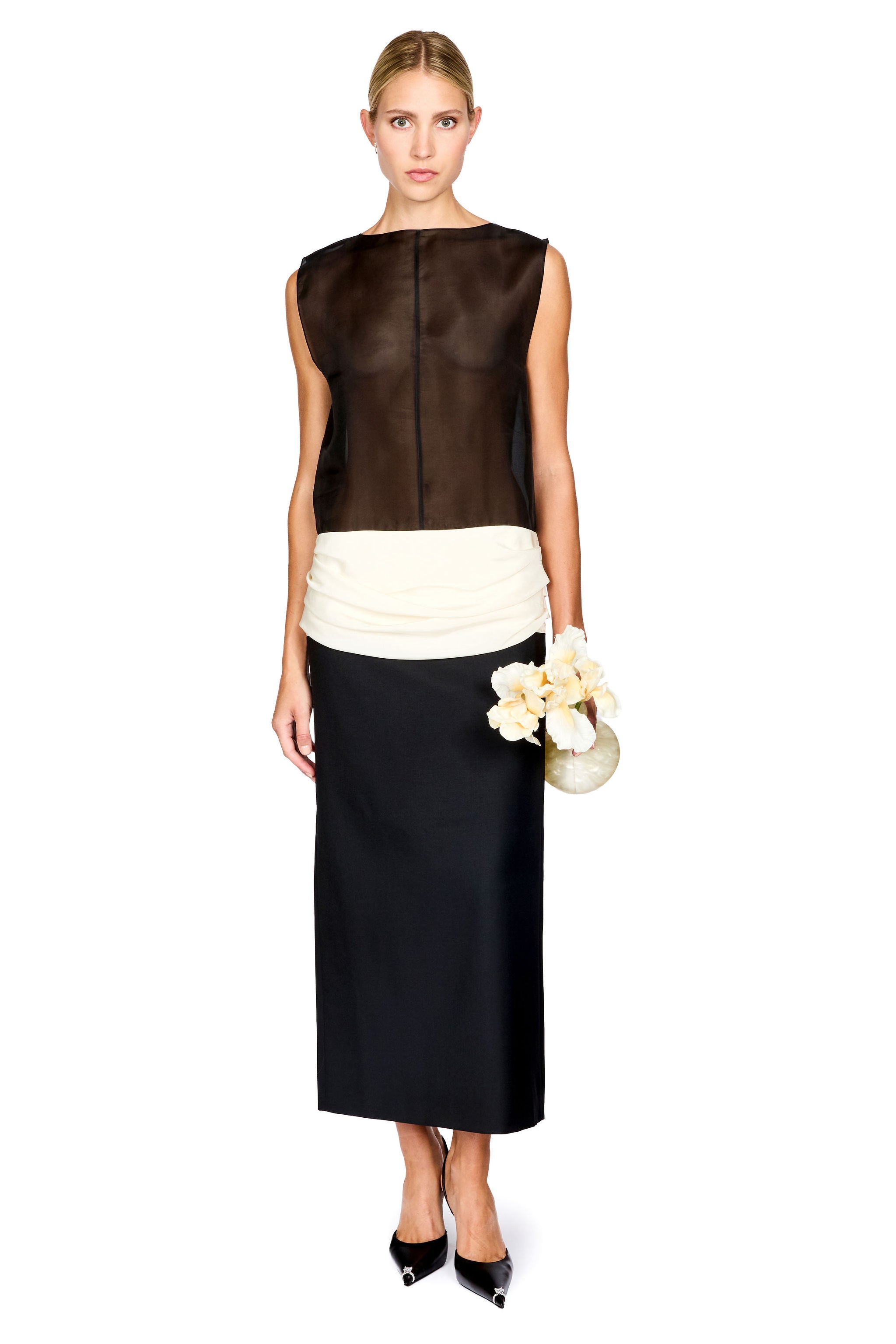 A woman standing in front of a white background wearing a black and white colorblocked long dress with a sheer top and white ruched waistband, holding a flower shaped clutch and black heels