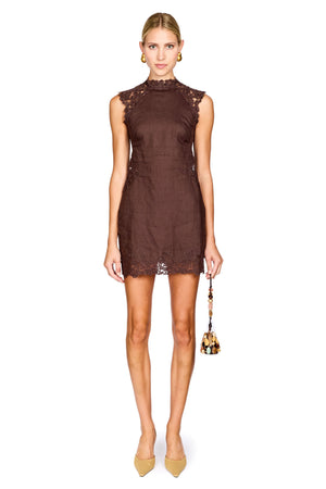 A model wearing a sleeveless, high-neck brown lace mini dress, holding a small gold and brown handbag, and wearing beige heels, set against a plain white background.