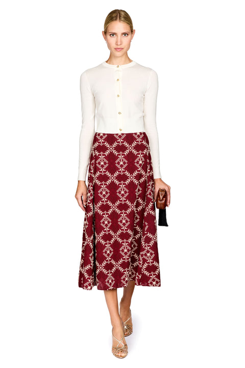 A model wearing a cream long-sleeve knit top and a burgundy midi skirt with a cream geometric pattern, holding a small black clutch against a plain white background.