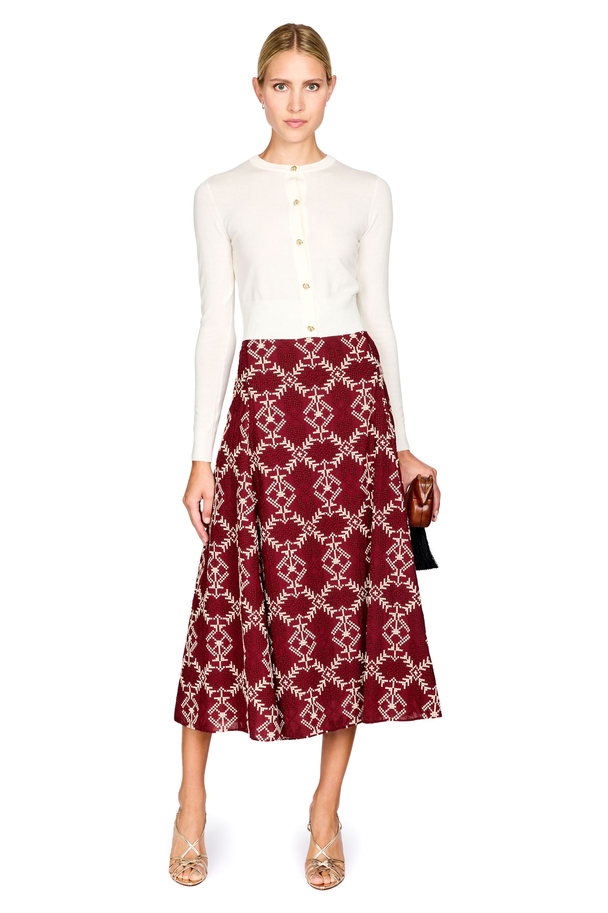 A model stands against a white studio background wearing a cream ribbed long-sleeve button-front top and a high-waisted, flared midi skirt with a burgundy and cream geometric pattern. She is wearing clear-strap high heels and holding a small black handbag.