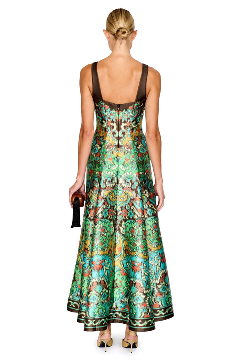 A woman viewed from the back wearing a sleeveless, floor-length dress with a rich green and turquoise paisley pattern, brown straps, and a dark brown back panel. She is holding a small black clutch against a solid white background.
