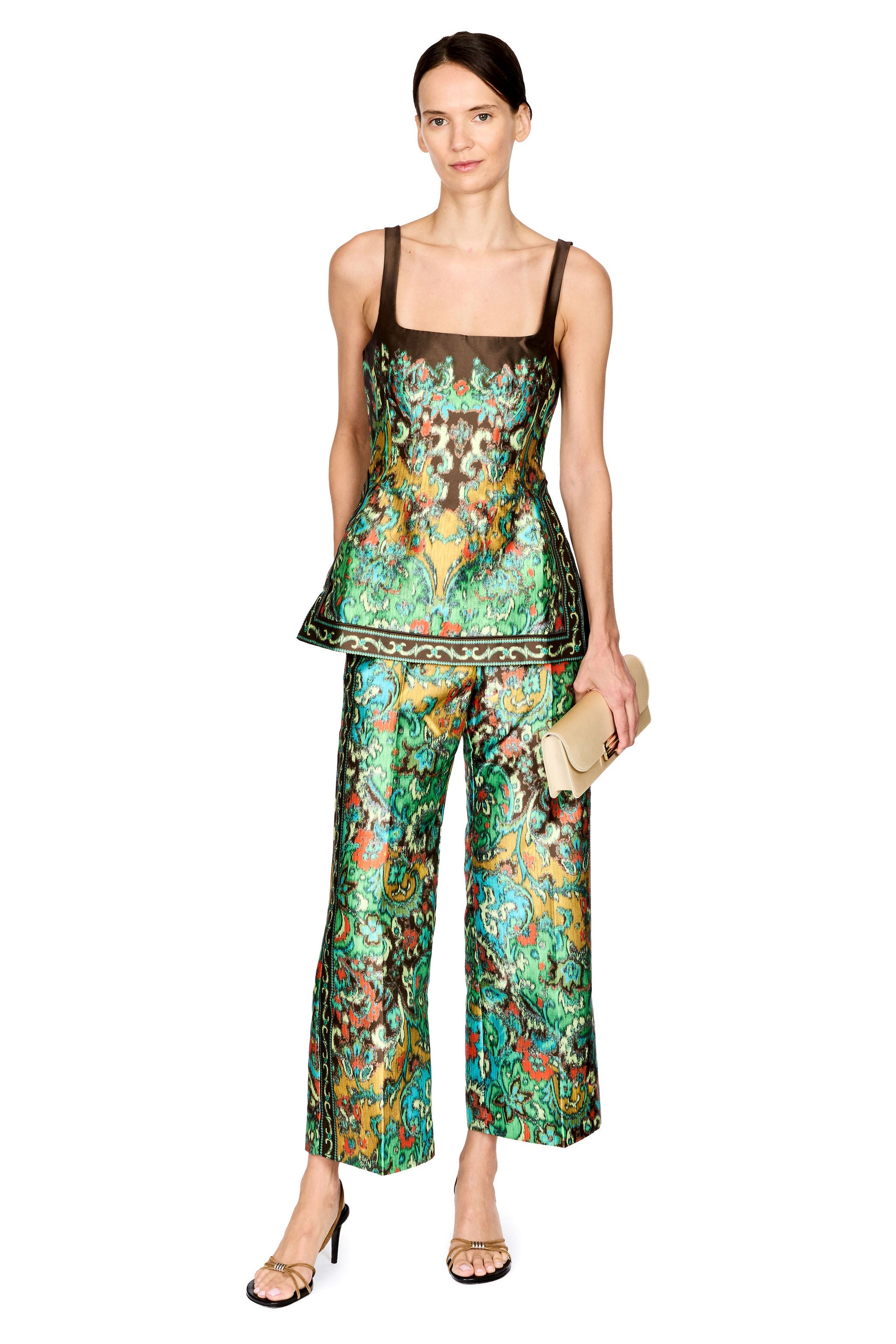 A model stands against a white background wearing a matching two-piece set consisting of a sleeveless peplum top and wide-leg pants, both featuring a vibrant green, blue, and orange floral pattern with a dark brown mesh underlayer. She is holding a small beige clutch and wearing gold sandals.
