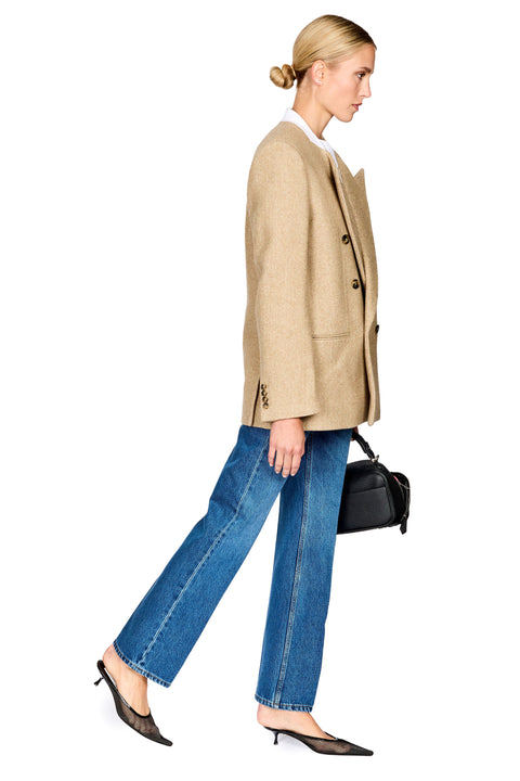 A woman in side profile wearing an oversized beige blazer, blue wide-leg jeans, black pointed-toe heels, and carrying a small black handbag against a plain white background.