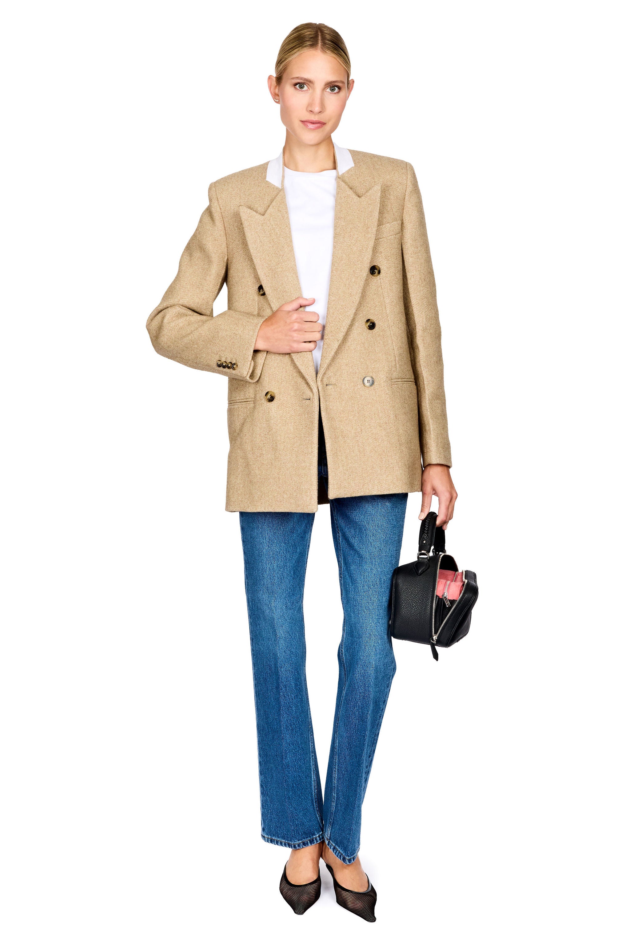 A woman modeling an oversized beige double-breasted blazer over a white top, paired with blue jeans, black pointed flats, and holding a small black handbag against a plain white background.