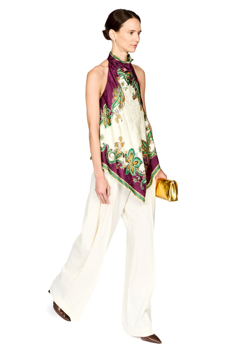 A model wearing a cream silk scarf with a purple border and green floral baroque pattern, styled as a sleeveless top over white wide-leg trousers, holding a gold clutch against a white background.