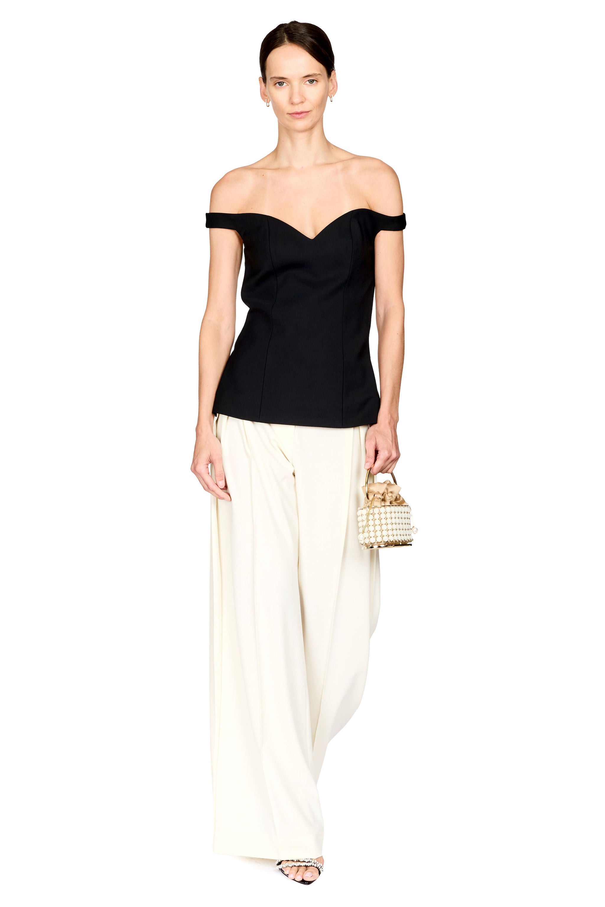 A model wearing a black off-the-shoulder top and cream wide-leg trousers, holding a small woven handbag against a plain white background.
