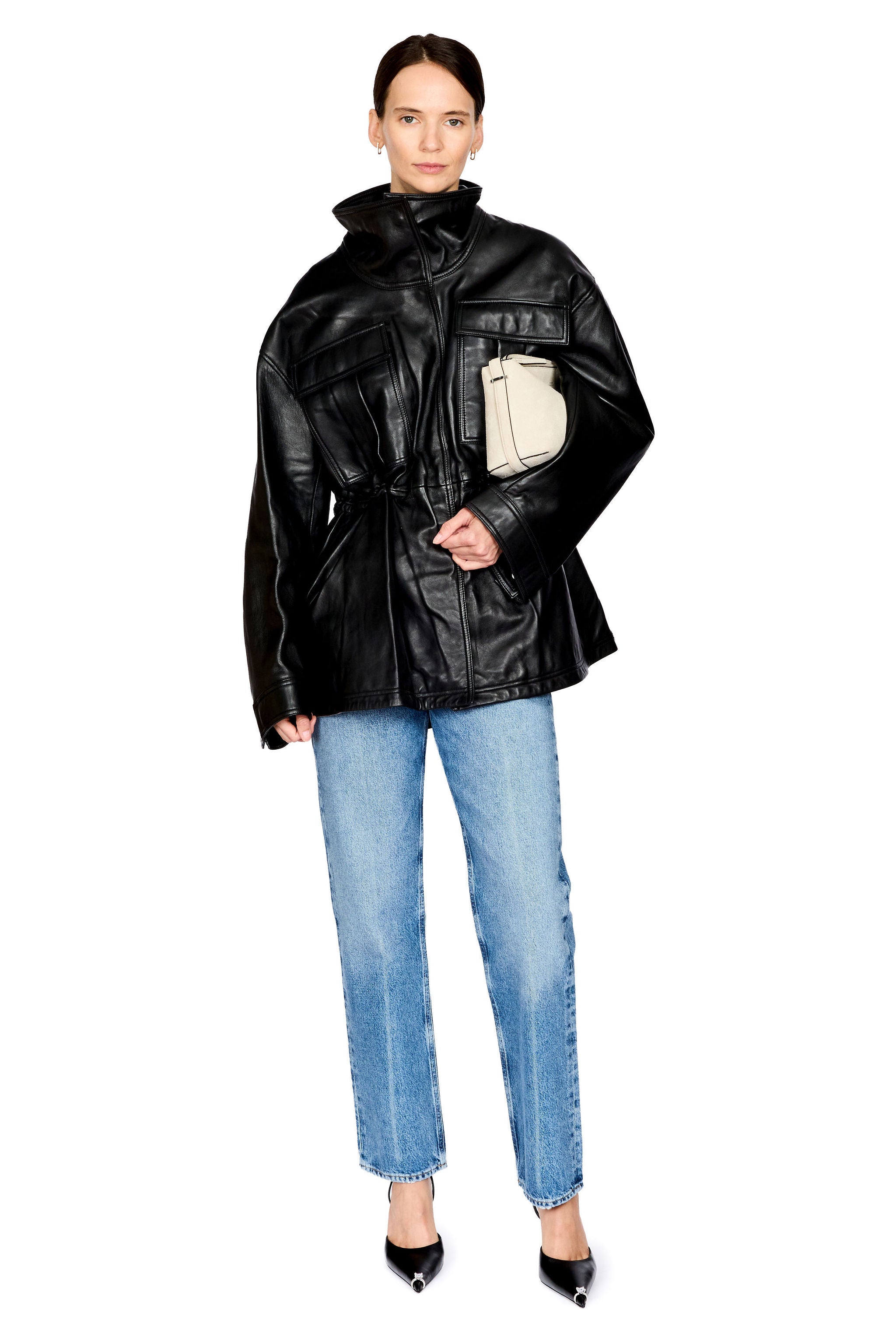 A model wearing a black leather jacket with a beige rectangular patch pocket, paired with light blue jeans and black pointed-toe heels, on a solid white background.