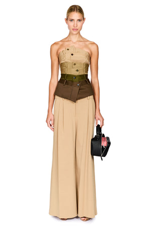 A model stands against a white studio background wearing a strapless beige and brown structured top with wide-leg beige pants, holding a small black handbag.