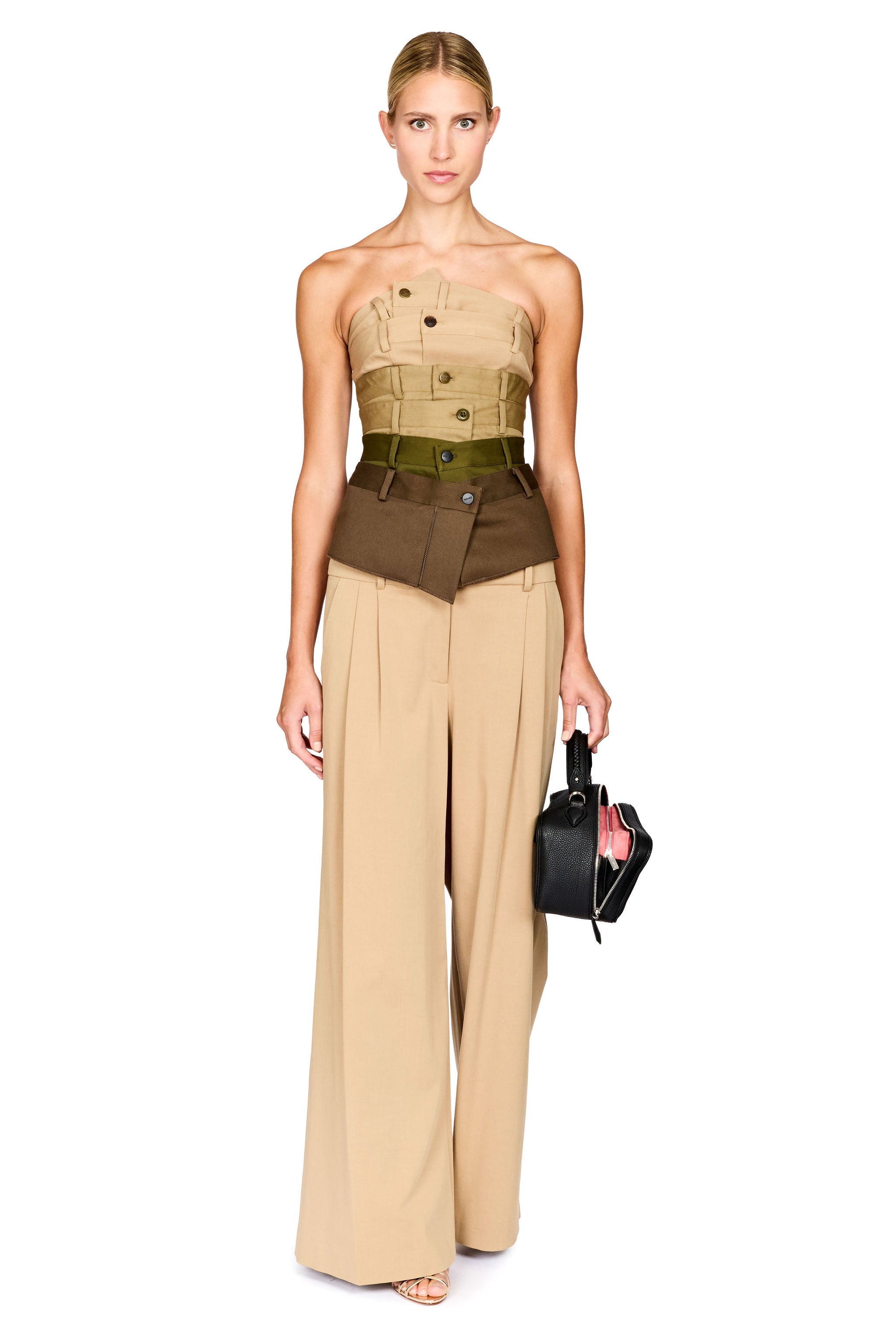 A model stands against a white background wearing a structured strapless top and wide-leg pants in beige, brown, and olive green, holding a small black handbag.