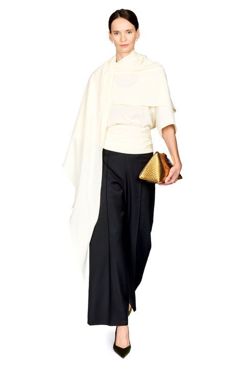 A model wearing a cream draped shawl over a cream top and black wide-leg trousers, holding a gold textured clutch against a solid white background.