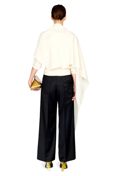 A model shown from the back wearing a cream draped cape-style top, black wide-leg trousers, and metallic gold heels, holding a gold clutch against a plain white background.