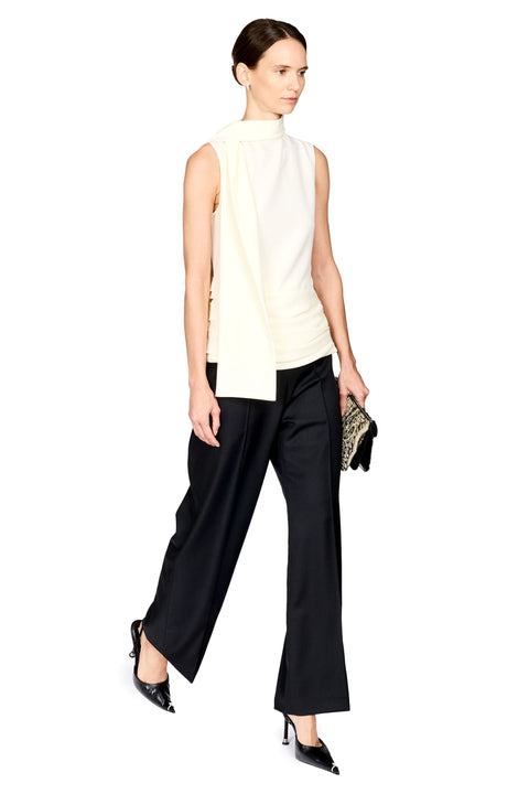 A model is shown from the back wearing a sleeveless cream mock-neck top and black wide-leg trousers, holding a small patterned clutch against a solid white background.