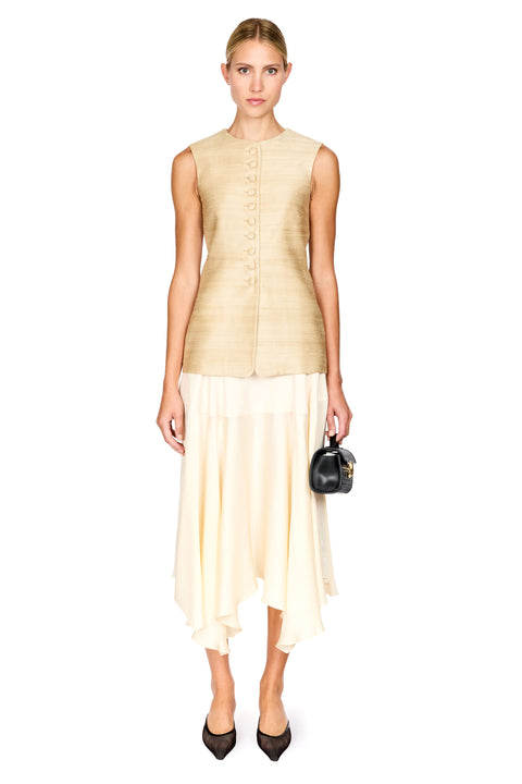 A model stands against a white studio background wearing a sleeveless beige textured top with front buttons and a flowing cream asymmetrical skirt, holding a small black handbag.