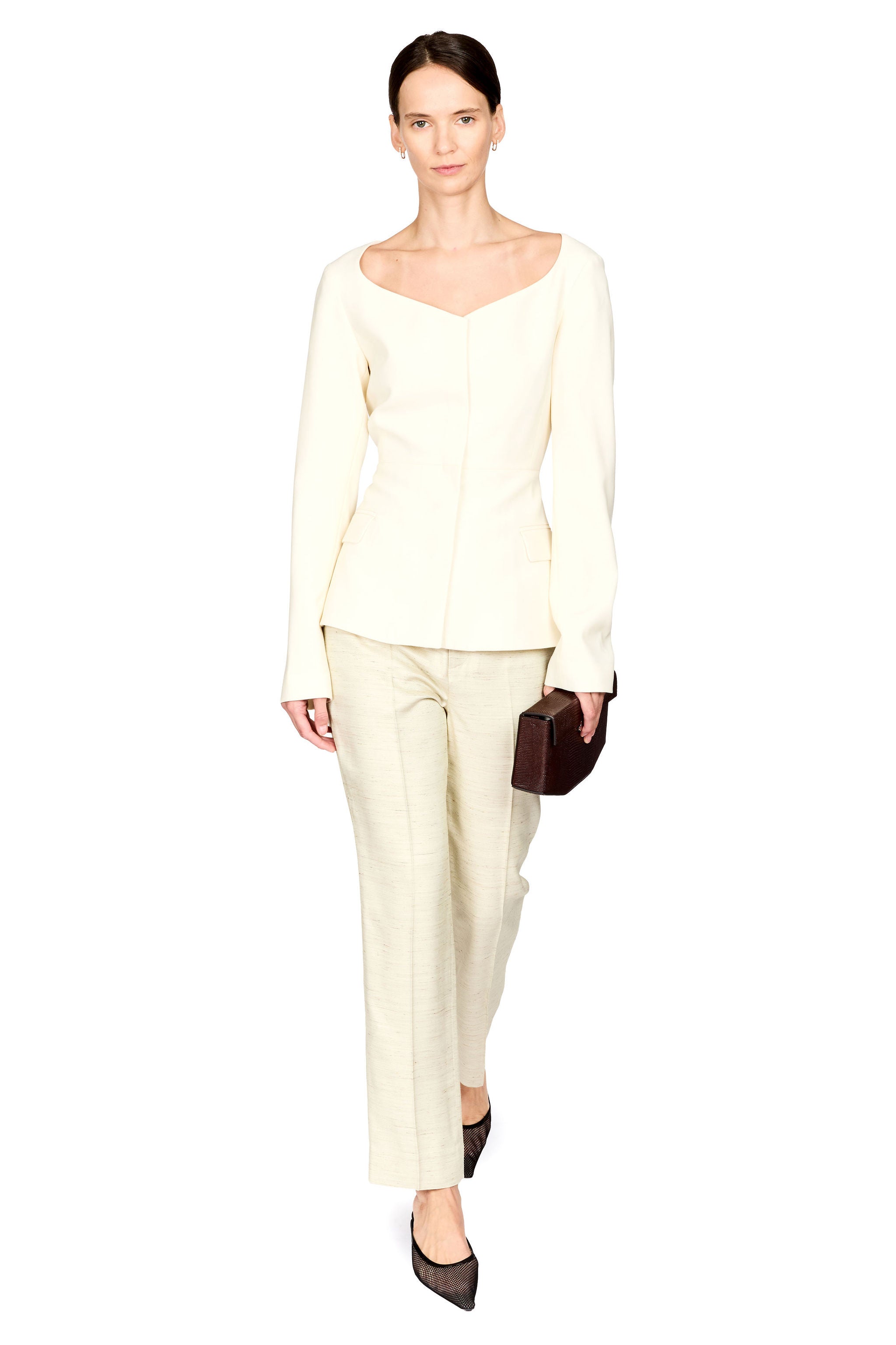 A model wearing a cream-colored two-piece suit consisting of a long-sleeve peplum-style top and matching trousers, holding a small dark brown clutch against a solid white background.
