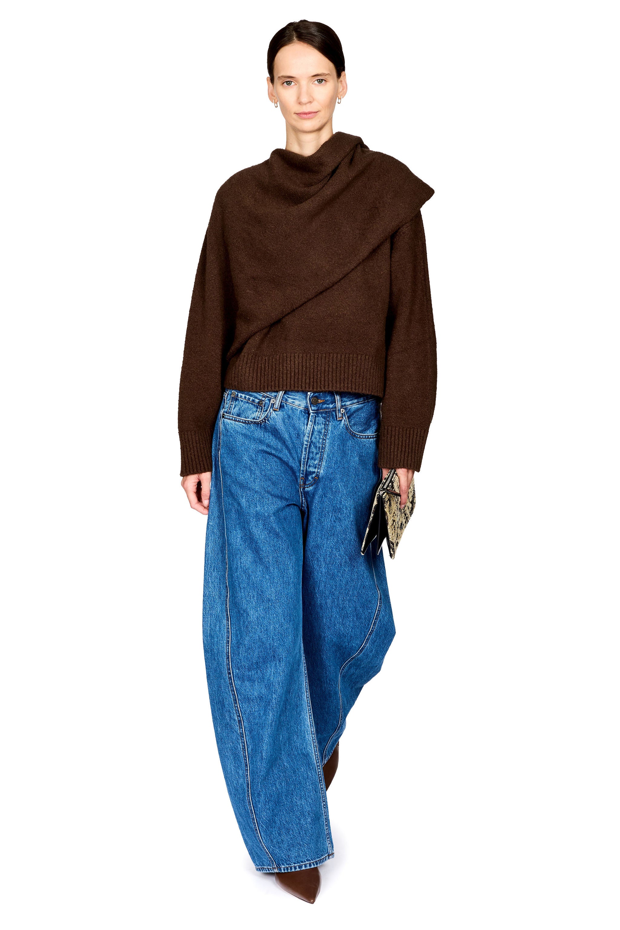 A model wearing a dark brown asymmetrical cowl-neck knit sweater, wide-leg blue denim jeans, and brown pointed-toe boots, holding a small patterned clutch against a plain white background.