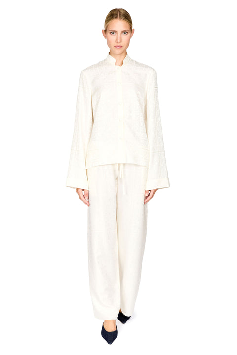 A woman modeling a matching cream-colored two-piece outfit consisting of a long-sleeve cropped jacket with a mandarin collar and wide-leg trousers, set against a plain white background.