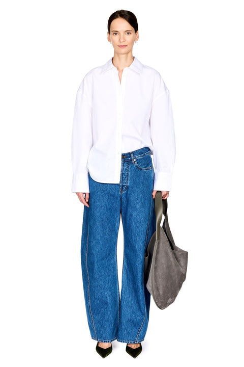 A model stands against a white studio background wearing an oversized white button-up shirt, wide-leg blue jeans, black pointed shoes, and holding a large gray tote bag.