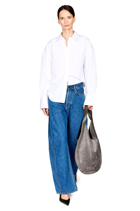 A model stands against a white studio background wearing an oversized white button-down shirt, wide-leg blue jeans, and black pointed shoes, while carrying a large gray suede shoulder bag.