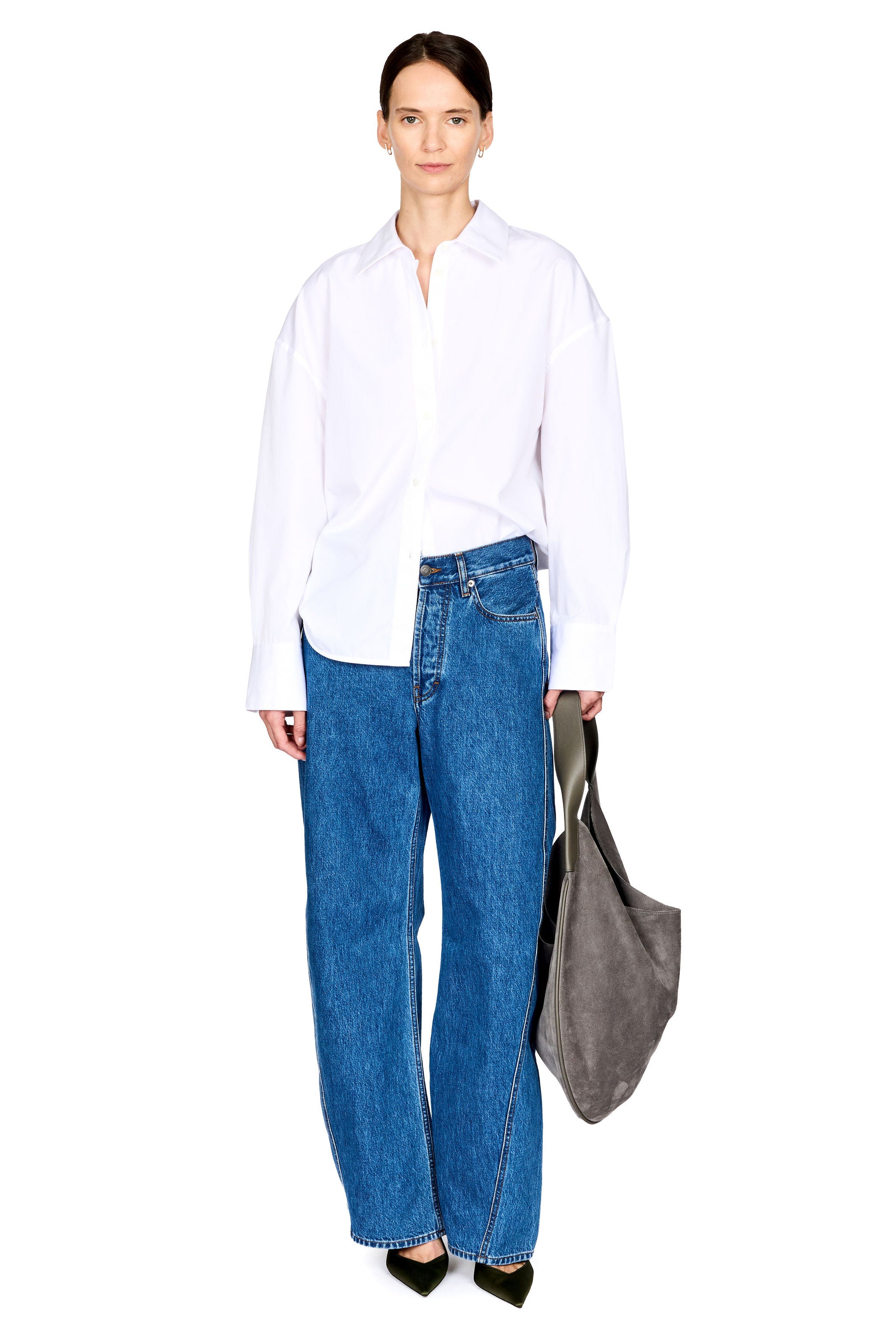A model stands against a white studio background wearing an oversized white button-up shirt, wide-leg blue jeans, black pointed shoes, and carrying a large gray tote bag.