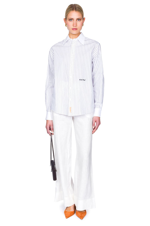 Woman wearing a striped shirt and white pants on a white background with one hand clutching bag.