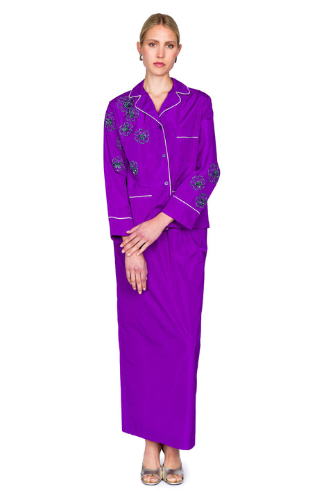 Woman wearing a purple button down pajama shirt with floral embellishments and a matching maxi skirt holding one wrist.