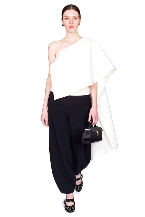 Woman wearing a white one-shoulder top and black pants clutching a black bag on a white background with one foot in front of the other.