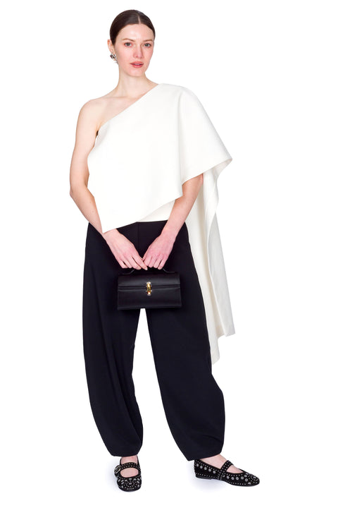 Woman wearing a white one-shoulder top and black pants clutching a black bag with both hands on a white background.