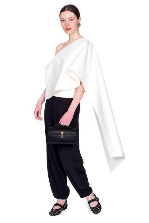 Woman wearing a white one-shoulder top and black pants clutching a black bag on a white background from the side view.