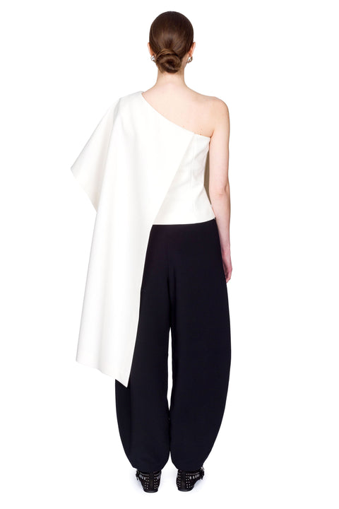 Woman wearing a white one-shoulder top and black pants clutching a black bag on a white background from the back.