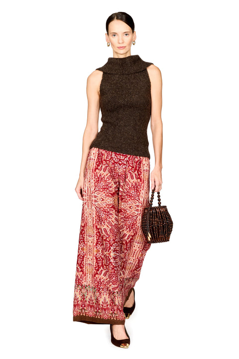 A model wearing a sleeveless dark brown mock-neck knit top and wide-leg red and beige patterned pants, holding a small dark woven handbag against a plain white background.