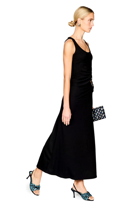 A model stands against a white background wearing a sleeveless black top and maxi skirt with a draped waist detail and a gold-toned ring accent. She is holding a small black patterned clutch and wearing blue patterned flat sandals.