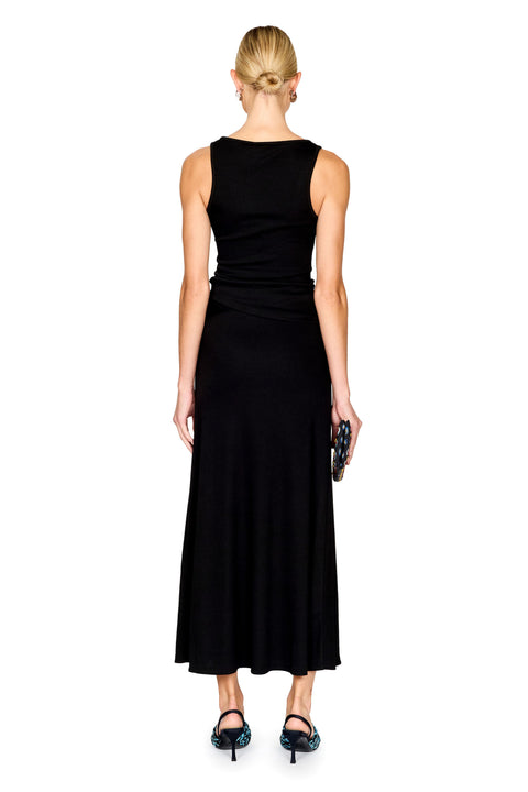 A model stands against a white background wearing a sleeveless black top and maxi skirt with a draped waist detail and a gold-toned ring accent. She is holding a small black patterned clutch and wearing blue patterned flat sandals.