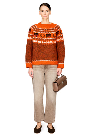 A model stands against a white studio background wearing an orange and brown patterned knit sweater, beige wide-leg jeans, dark brown flats, and holding a brown handbag.