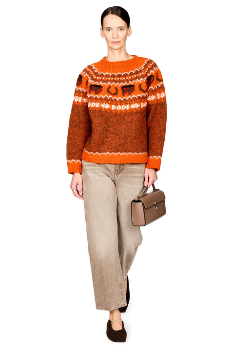 A woman modeling a bright orange and brown patterned knit sweater, paired with beige wide-leg jeans, dark brown flats, and holding a small brown handbag against a plain white background.