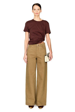 A model stands against a white background wearing a dark brown short-sleeve t-shirt with a twisted front detail, paired with high-waisted, wide-leg tan trousers and holding a small clutch.