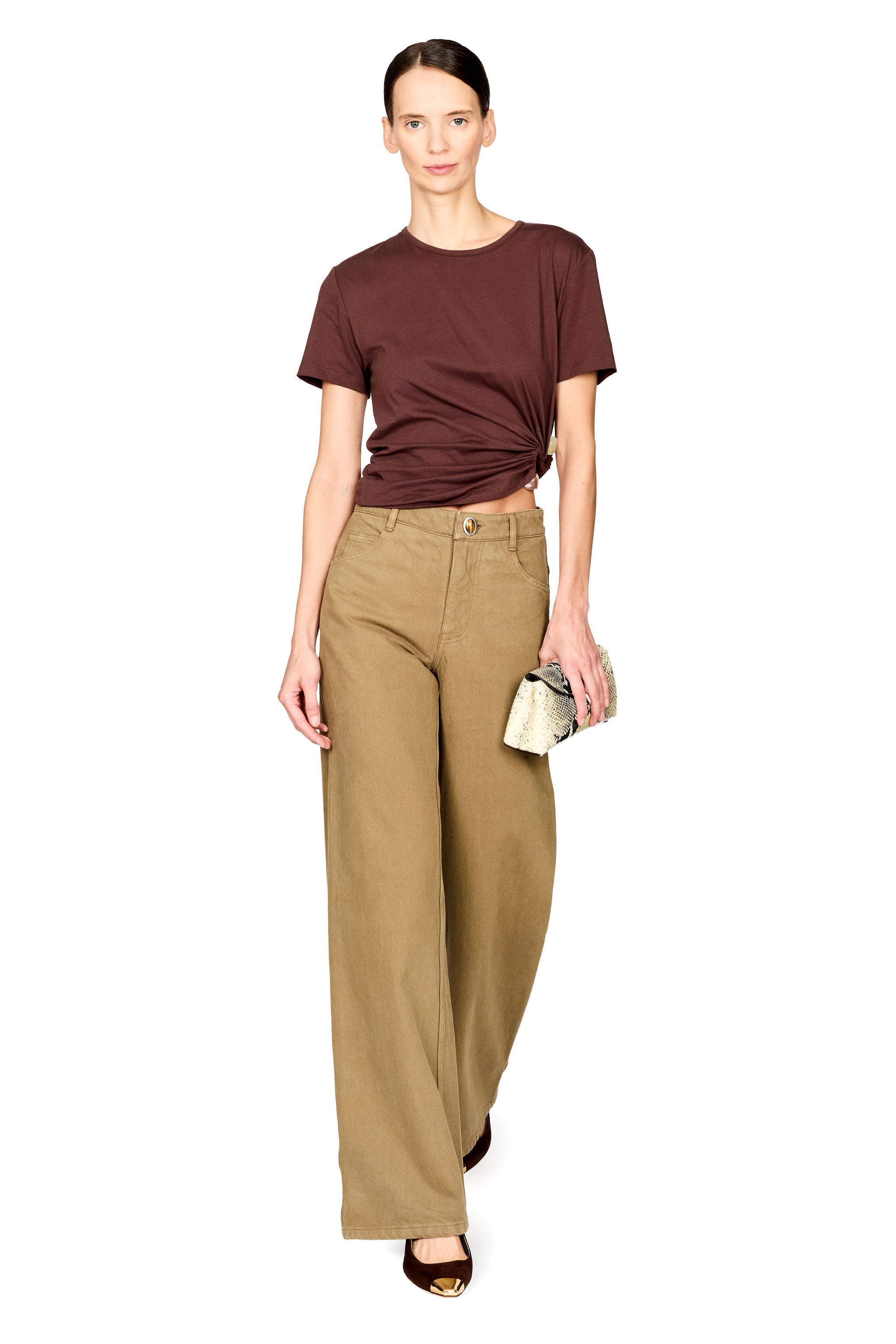 A model wearing a dark brown short-sleeve t-shirt with a twisted hem, paired with high-waisted, wide-leg khaki trousers and holding a small metallic clutch, set against a plain white background.