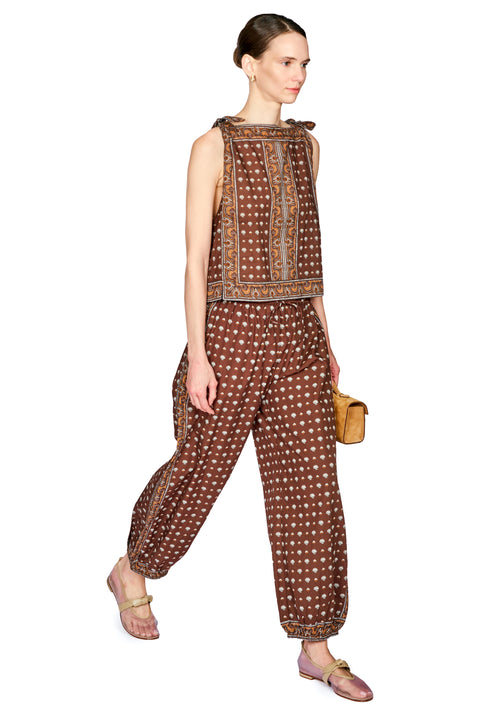 A model wearing a sleeveless brown jumpsuit with a white polka dot pattern and a decorative embroidered border, paired with pinkish-brown flat sandals and holding a small tan handbag, set against a plain white background.