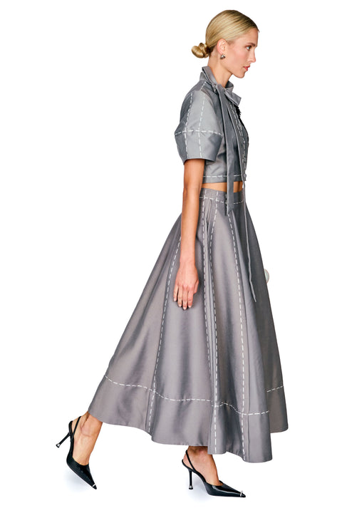 A woman modeling a gray two-piece outfit consisting of a short-sleeve cropped shirt with a tie collar and a matching high-waisted flared skirt, both featuring white contrast stitching details, paired with black pointed-toe heels against a white background.