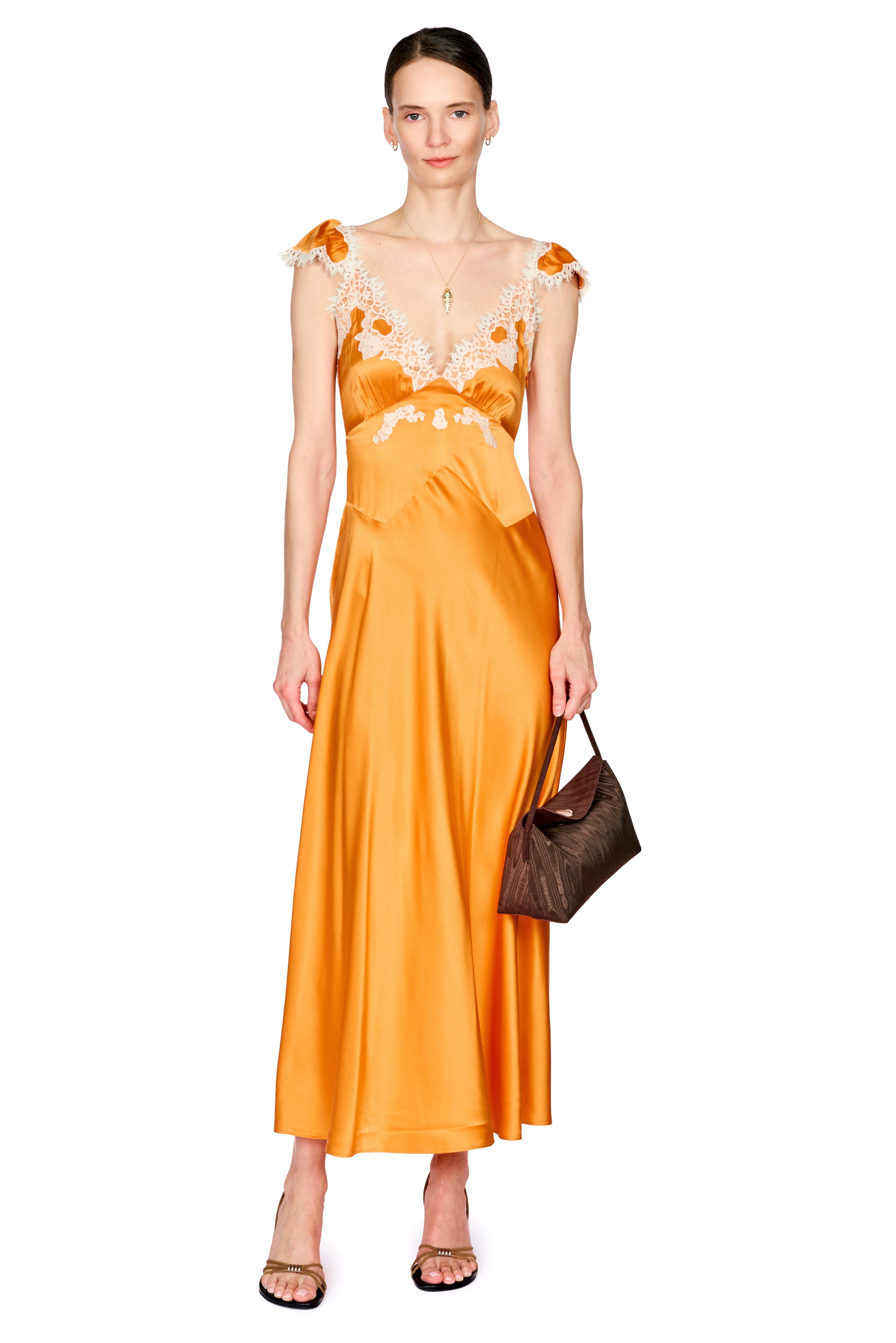 A woman modeling a bright orange satin maxi dress with floral lace embroidery on the bodice and shoulders, holding a brown handbag against a plain white background.