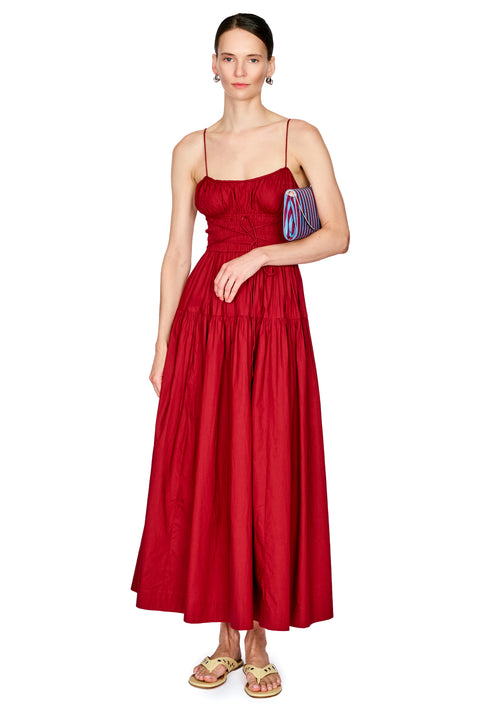 A woman modeling a red maxi dress with thin spaghetti straps, a gathered ruched bodice, and a tiered skirt, paired with gold sandals and a small striped clutch, on a solid white background.