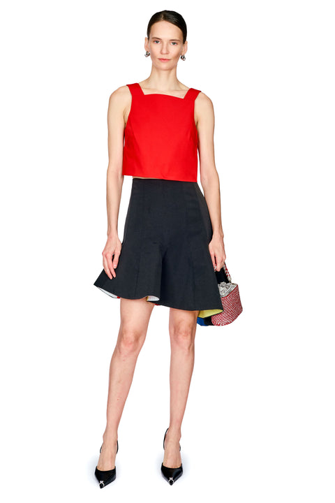 A model stands against a white background wearing a sleeveless red top and a black flared skirt, holding a small patterned handbag.