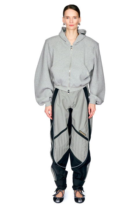 A model wearing a light gray zip-up hoodie and matching gray and black athletic pants, standing against a plain white background.