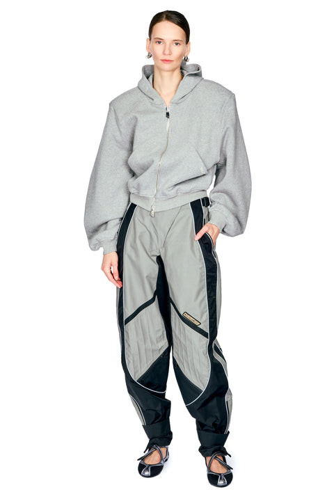 A model wearing a light gray cropped zip-up hoodie and matching gray jogger pants with black side panels and black zipper details, paired with black flats, against a plain white background.