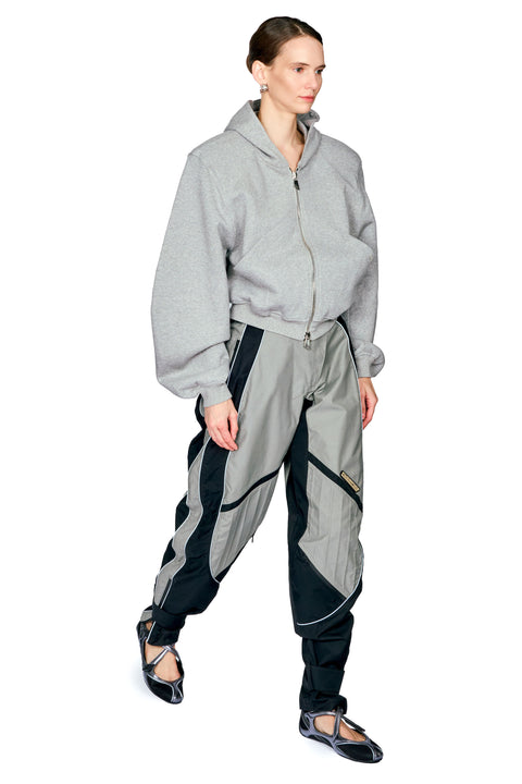A model wearing a light gray zip-up hoodie and matching gray and black track pants, standing against a plain white background.